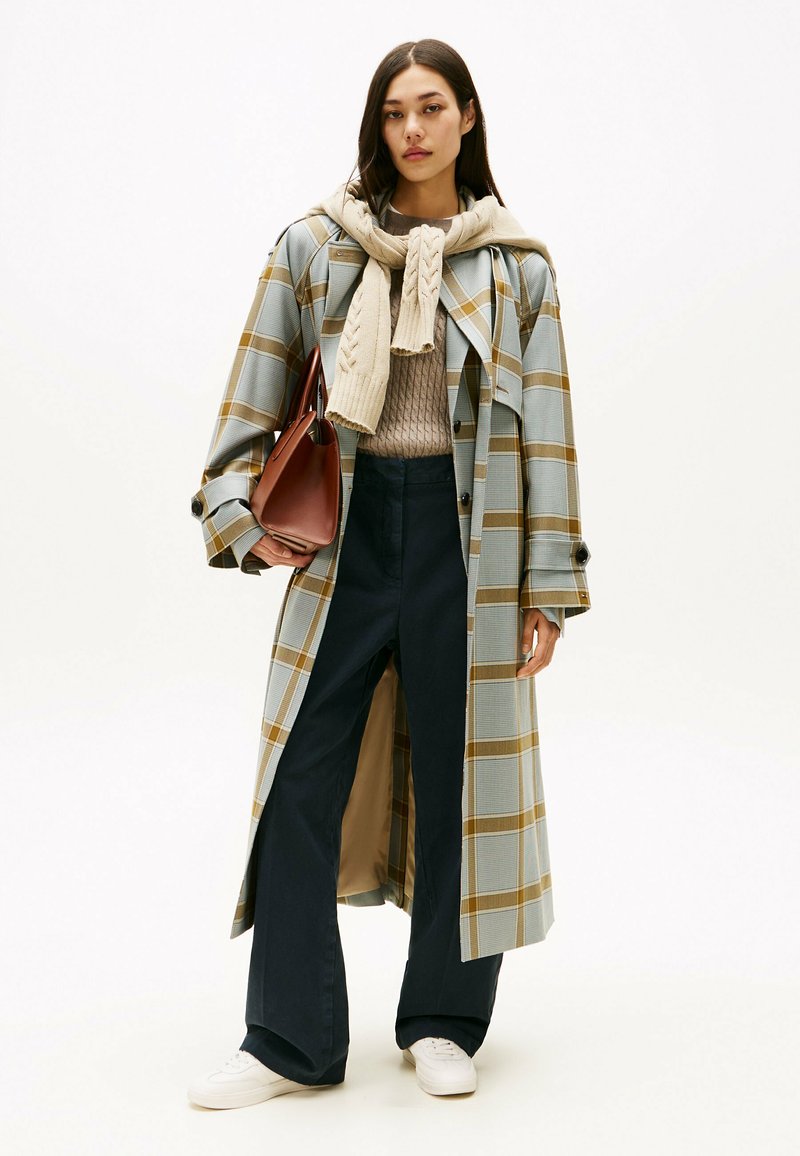 Abrigo largo de cuadros en azul y amarillo, combinado con un suéter beige de punto trenzado y pantalones anchos oscuros. Bolso marrón y zapatillas blancas.