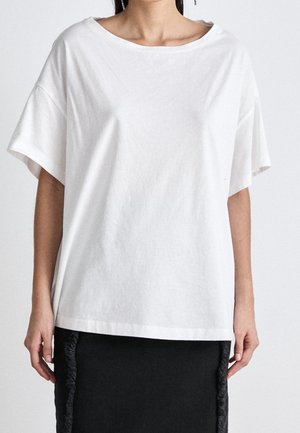 Mujer con una blusa blanca de manga corta y holgada combinada con una falda negra con detalles de flecos laterales, posando frente a un fondo liso.