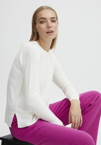 Pull en tricot blanc à manches longues et fentes sur les côtés, associé à un pantalon rose vif. Texture lisse et design simple.