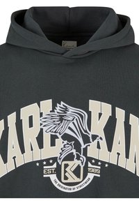 Sweat à capuche foncé avec un grand texte beige "KARL KANI", un graphisme d'aigle et un emblème "EST. 1989 The Originator of Streetwear" sur le devant.