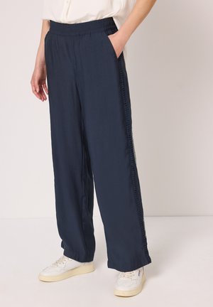 Pantalon ample bleu marine avec ceinture élastique et détails en dentelle sur les côtés, porté avec des baskets blanches et une chemise blanche rentrée.