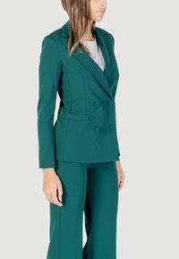 Modelo con un elegante blazer verde y pantalones a juego, mostrando un conjunto moderno y profesional frente a un fondo neutro.