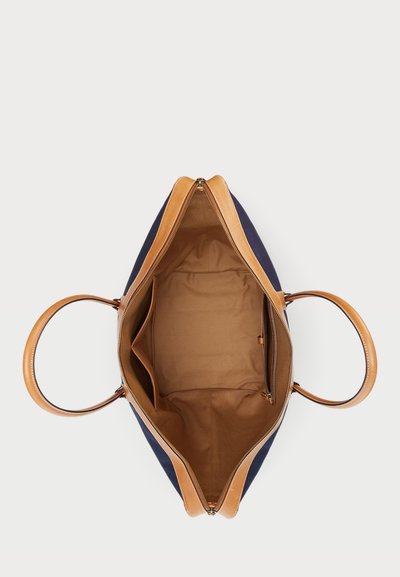 Saco tote de lona azul-marinho com alças de couro castanho e fecho de zipper. O interior apresenta um forro de tecido castanho com dois bolsos e um compartimento com fecho de zipper.