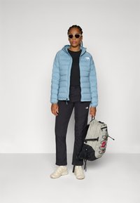 Light blue puffy jacket, black shirt, black pants, beige sneakers; carrying a gray-and-black backpack with red accents, on a neutral background.