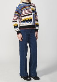 Maglione lavorato a maglia multicolore con motivi geometrici in blu, nero, giallo e cremisi. Abbinato a jeans a gamba larga blu scuro e scarpe nere.