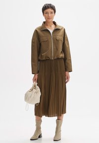 Veste zippée vert olive, jupe midi plissée marron, sac beige à cordon et bottines beige. Texture lisse et design décontracté.