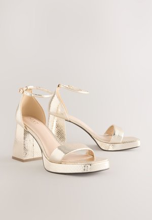 Sandales à talons hauts métalliques dorées avec un motif reptile texturé, bout ouvert, bride autour de la cheville, et talon épais bloc. Design à plateforme plate.