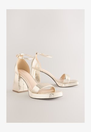 Sandales à talons hauts métalliques dorées avec un motif reptile texturé, bout ouvert, bride autour de la cheville, et talon épais bloc. Design à plateforme plate.