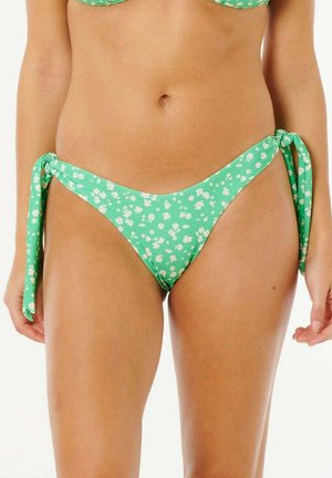 Braguita de bikini verde con patrón floral blanco, atada con lazos en ambas caderas, usada por una persona de pie contra un fondo liso.
