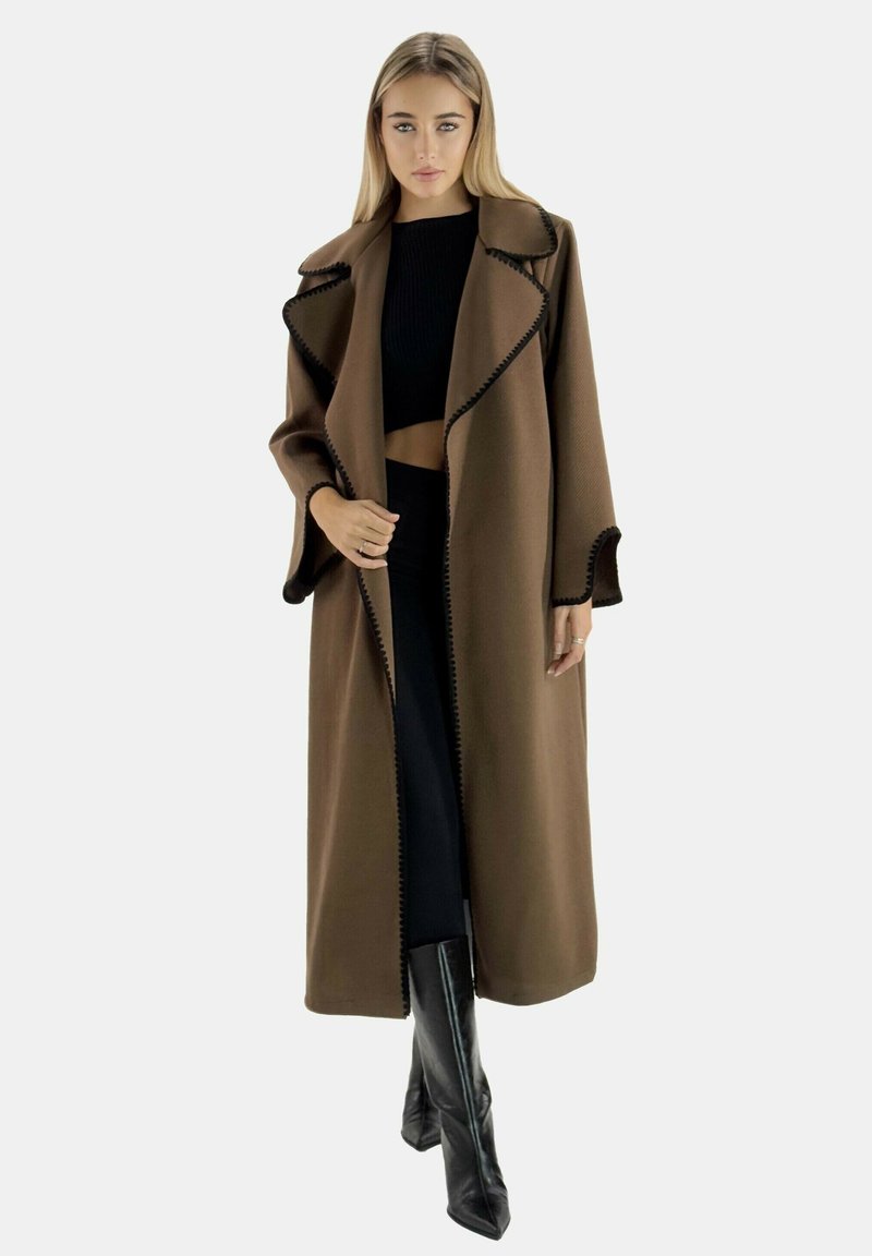 Cappotto lungo marrone con ampie rever, cuciture nere e design aperto. Indossato sopra un crop top nero e una gonna lunga, con stivali neri fino al ginocchio.