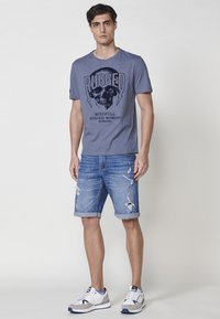 Mėlyna medvilninė marškinė su juodu grafiniu spaudiniu, derinami su suplėšytais džinsiniais šortais. Pilki ir balti sportiniai bateliai užbaigia kasdienį įvaizdį.