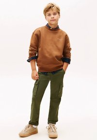 Felpa marrone con colletto rotondo, pantaloni cargo verdi e sneakers beige. Camicia a quadri visibile ai polsini, con due tasche cargo sui pantaloni.