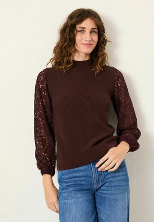 Braun geripptes Strickoberteil mit spitzen Ballonärmeln, ausgestattet mit elastischen Manschetten. Kombiniert mit hellblauen Jeans. Einfarbiger Hintergrund.