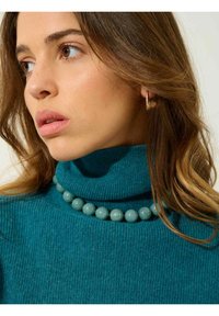 Teal ribbad turtleneck-tröja med en åtsittande krage och ett runt, glansigt turkos pärlhalsband. Bärs med små hoopörhängen.