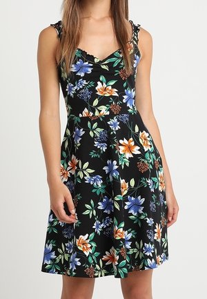 Robe noire sans manches avec motif floral bleu, orange et blanc, taille cintrée et jupe longueur genou portée par une femme aux longs cheveux bruns.