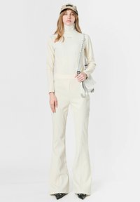 Mono ajustado de color crema con piernas acampanadas, cuello alto y detalles visibles de costuras. Accesorio: gorra beige con un logotipo.