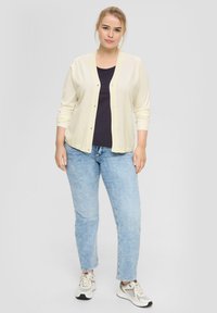 Hellgelbe Strickjacke mit Knopfleiste, über einem dunklen Oberteil getragen, kombiniert mit hellblauen Jeans und grauen Sneakers.