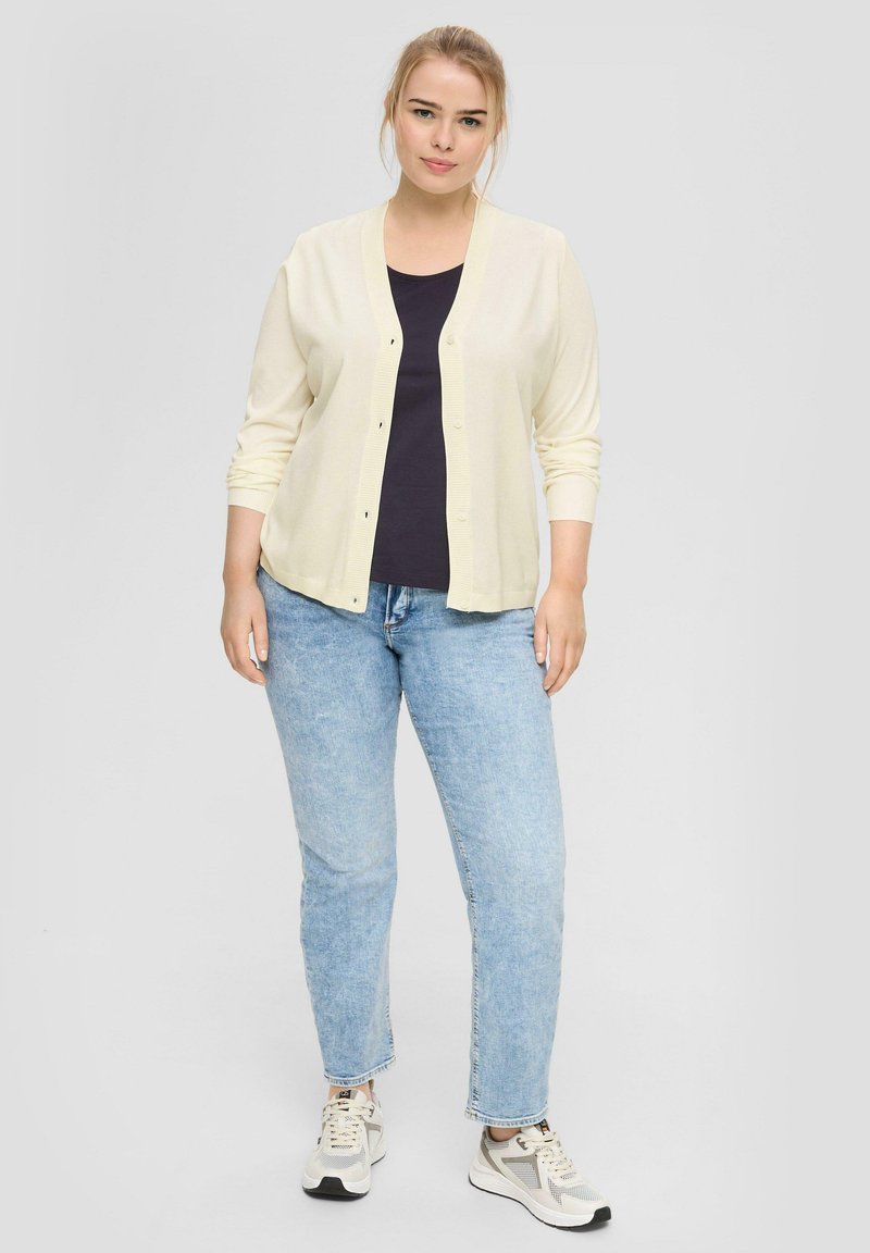 Hellgelbe Strickjacke mit Knopfleiste, über einem dunklen Oberteil getragen, kombiniert mit hellblauen Jeans und grauen Sneakers.