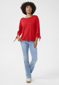 Red long-sleeve top with a wide neckline, elasticated hem, and tie details on the sleeves, paired with light blue jeans and sneakers.