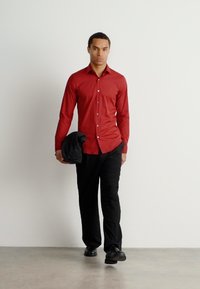 Červená košile s knoflíky, slim fit, ve spojení s černými kalhotami. Model drží složenou tmavou bundu, má na sobě černé boty, na neutrálním pozadí.