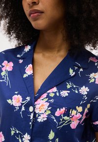 Γυναίκα που φοράει πουκάμισο σε σκούρο μπλε με λουλούδια σε ροζ και λευκό και κεντημένο το γράμμα "RL" στην τσέπη στο στήθος.