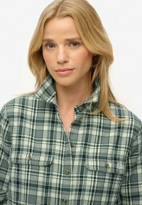 Blonde Frau mit blauen Augen, die ein grün-weiß kariertes Hemd mit Knöpfen und Brusttaschen trägt, vor einem einfarbigen Hintergrund.