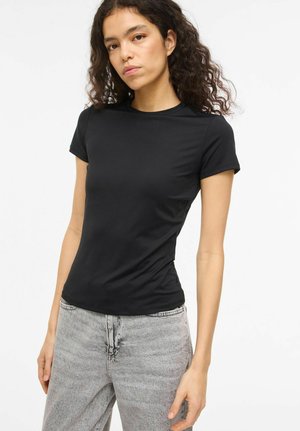 Mlada ženska s kodrastimi lasmi, oblečena v pripeto črno majico s kratkimi rokavi in svetlo sive oprane kavbojke, na belem ozadju.