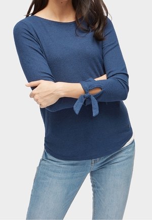 Woman wearing a blue long-sleeve top with tied cuffs and light blue jeans, arms crossed at waist, against a plain background.
