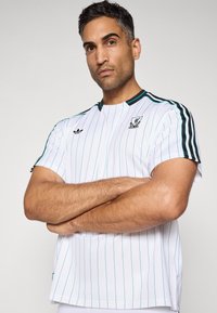 Mand med armene folderet iført en hvid, stribet Adidas sportsjersey med Liverpool FC-logo på en ensfarvet baggrund.
