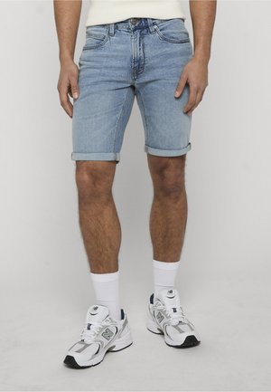 Mężczyzna stojący w jasnoniebieskich podwiniętych szortach dżinsowych, białych skarpetkach do kostek oraz białych butach sportowych New Balance.