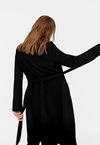 Abrigo largo negro con cinturón, que presenta una textura suave y un diseño de frente abierto, mostrando un acabado liso y detalles de mangas a medida.