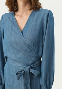 Robe portefeuille en denim bleu clair avec des manches longues, un col en V et une taille nouée. Présente une texture douce et des rayures verticales subtiles.