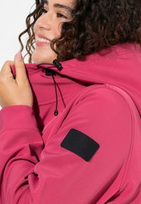 Rosa huva jacka gjord av mjukt material. Har dragsko stängning vid halsen och en läderlogotyp på vänster axel. Slät textur.