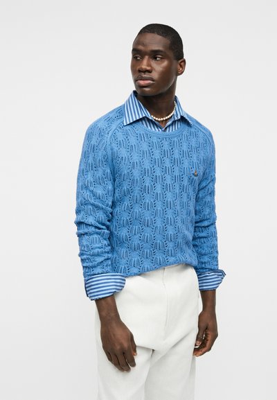 Άνδρας που φοράει μπλε πλεκτό πουλόβερ με σχέδια πάνω από μπλε ριγωτό πουκάμισο με γυρισμένα μανίκια, λευκό παντελόνι και περιδέραιο με πέρλες, κοιτάζοντας δεξιά.