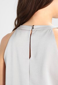 Ljust grå ärmlös topp med en nyckelhålsdetalj i ryggen, liten metallknapp i stängning och ett subtilt, texturerat tygmönster.