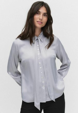Blouse gris clair avec col, manches longues et boutons sur le devant. Présente un détail plissé et une texture satinée lisse.