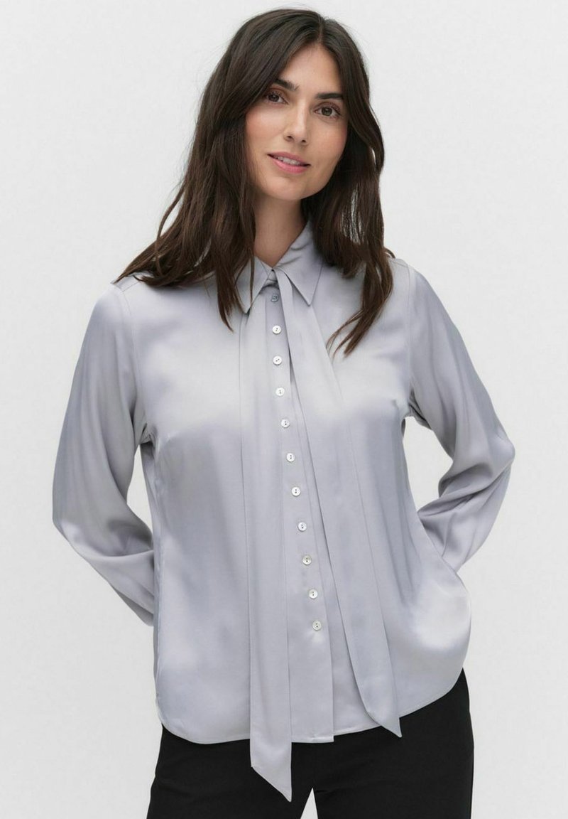 Lichtgrijze blouse met een kraag, lange mouwen en knopen aan de voorkant. Heeft een geplisseerd detail en een soepele satijnen textuur.