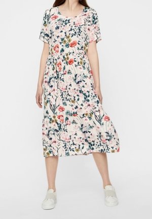 Nainen valkoisessa, monivärisellä kukkakuviolla varustetussa midi-mekossa, jossa on lyhyet hihat, valkoiset liukusovituskengät ja jalat hieman erillään seisten.