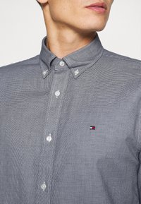 Camicia a bottoni blu navy con un piccolo motivo a pois bianchi, dotata di colletto e orlo arrotondato. Piccolo logo in rosso, bianco e blu sul petto.