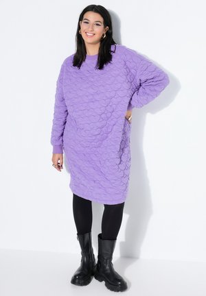 Smilende kvinde med langt mørkt hår iført tekstureret lavendelfarvet sweaterkjole, sorte leggings og sorte chunky støvler, stående mod en hvid væg.