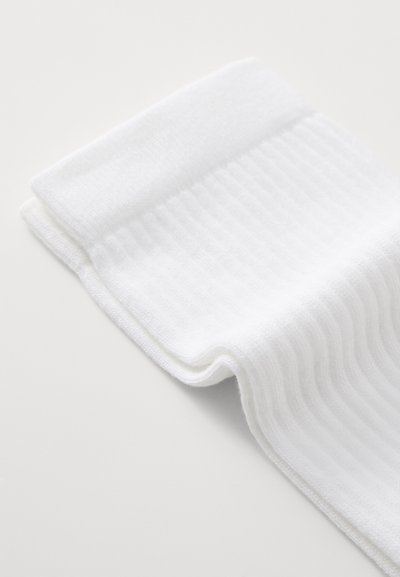 Hvidt sneakersok med ribbet tekstur, der har et blødt stof og en ombukket top. Sokken er foldet, hvilket fremhæver dens glatte overflade.