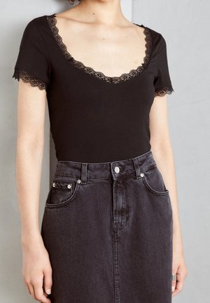 Vrouw draagt een zwart topje met korte mouwen en kantafwerking bij de halslijn en mouwen, gecombineerd met een high-waisted donkerblauwe denim rok.