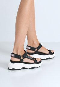 Svarta textilsandaler med justerbara remmar, med en kraftig vit sula och strukturerade detaljer, utformade för komfort och stabilitet.