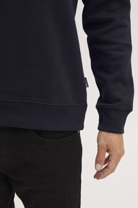 Sudadera azul marino con puños y dobladillo acanalados, hecha de material suave, mostrada con una mano descansando cerca de un bolsillo negro.