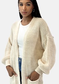 Beige gebreide cardigan met open voorkant, oversized mouwen en zijzakken, gedragen over een witte geribbelde top en blauwe jeans.