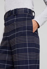 Marineblauwe geruite broek met een getailleerde pasvorm, zijzakken, gevoerde tailleband en afwisselend dunne witte en beige strepen in een ruitpatroon.