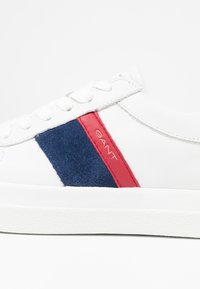 Białe skórzane sneakersy z niebieską zamszową wstawką i czerwoną paskiem akcentowym. Płaska biała gumowa podeszwa. Wytłoczone "GANT" na czerwonej części.