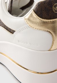 Nærbilde av en hvit sneakers med gull- og brunt aksenter, med preget "Michael Kors" merkevare på sidepanelet.
