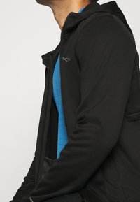 Man i en svart zip-up jacka över en blå skjorta, visad från nacke ner till mitten av torso i en närbild från sidan.