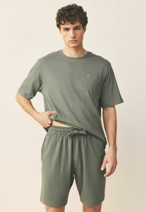 Junger Mann trägt ein olivgrünes kurzärmeliges Hemd und passende Shorts mit Kordelzug, hebt das Hemd leicht mit einer Hand an.